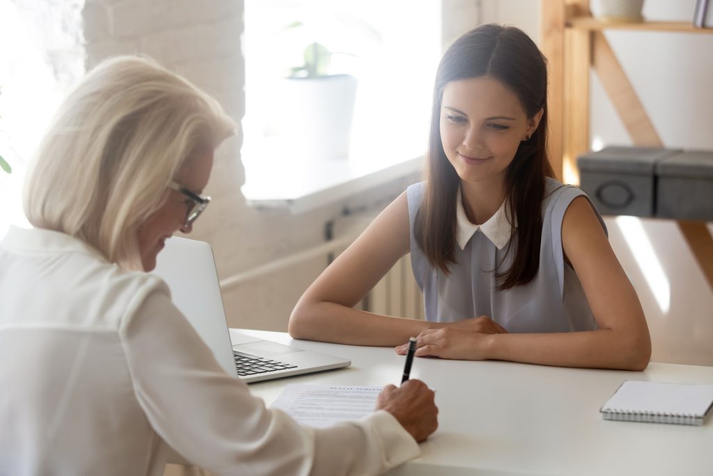 Jonge vrouw kijkt naar andere vrouw die een pen in de hand heeft en schrijft.